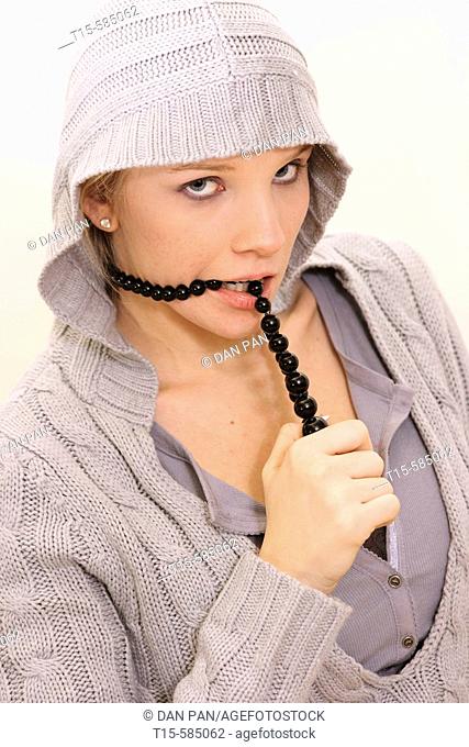 young woman dressed in a blue gray hooded sweater wearing a black beaded necklace and biting on the necklace looking playful or bored