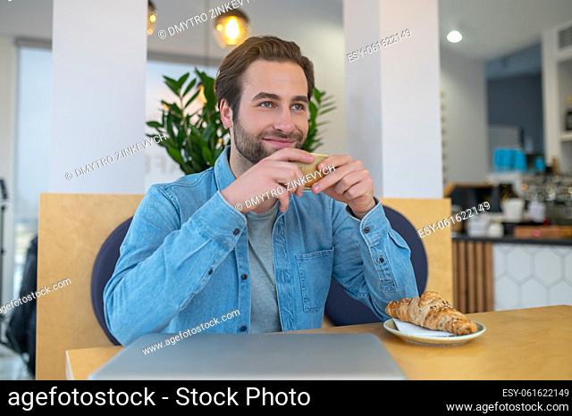 Break. Smiling young adult man with coffee looking away thoughtfully sitting at table with croissant and laptop in cafe