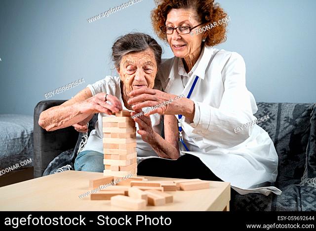 Jenga game. Theme is dementia, aging and games for old people. Caucasian senior woman builds tower of wooden blocks with the help of a doctor as part of a...