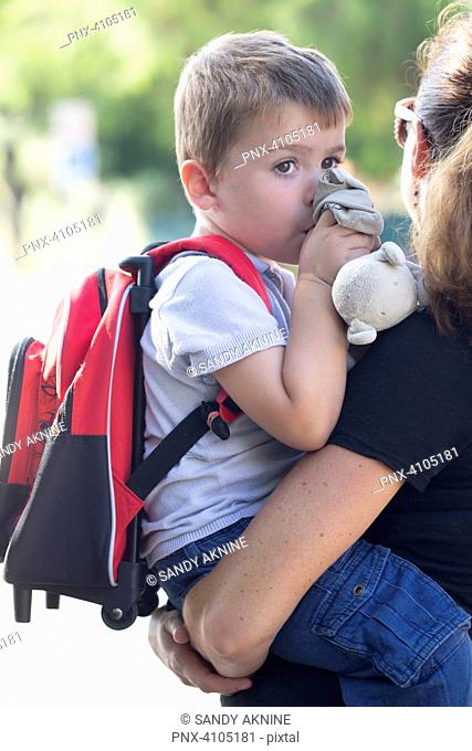 Portrait of a sad little boy in his mother's arms with his schoolbag and his thumb and his cuddly toy on his way out of school