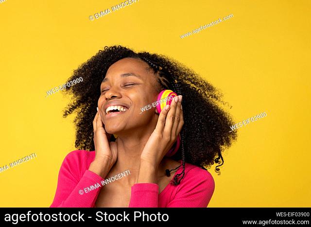 Happy young woman with eyes closed enjoying music through wireless headphones against yellow back ground