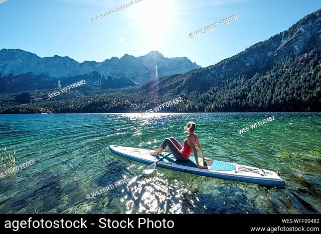 Germany, Bavaria, Garmisch Partenkirchen, Young woman sitting on stand up paddle board on Lake Eibsee and looking at Zugspitze Mountain
