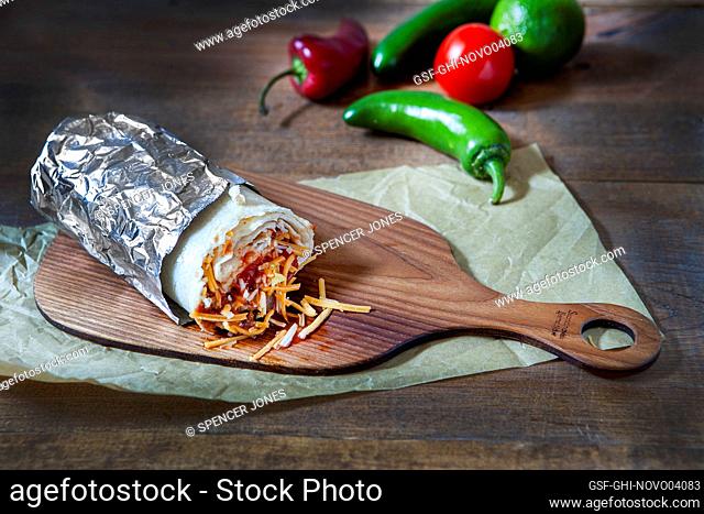 Partially eaten burrito wrapped in aluminum foil on wood board near red and green peppers and tomato