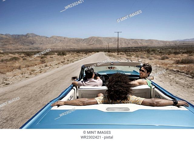 Three young people in a pale blue convertible car, driving on the open road across a flat dry plain