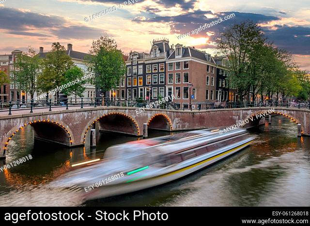 Netherlands. Summer sunset on the canals of Amsterdam. A tourist boat sails between the bridges. Traditional houses and bicycles on the waterfront