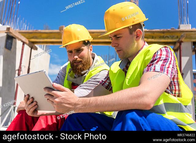 Side view of two workers, reading online information or watching a video on a tablet PC during break at work on the construction site of a contemporary building