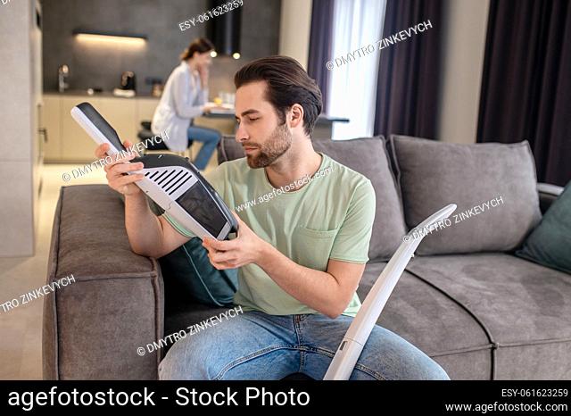 Interesting here. Attentive young bearded man examining detail of vacuum cleaner in hands sitting on sofa and woman behind