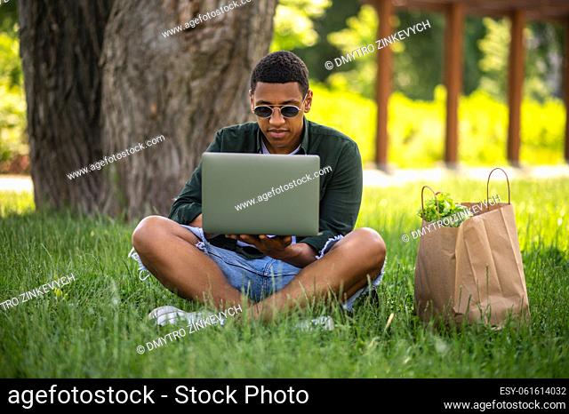 Time in nature. Dark-skinned guy in sunglasses looking into laptop sitting on grass in shade of tree in park