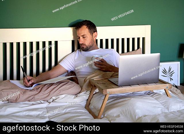 Man with coffee cup writing in book while working on bed at home