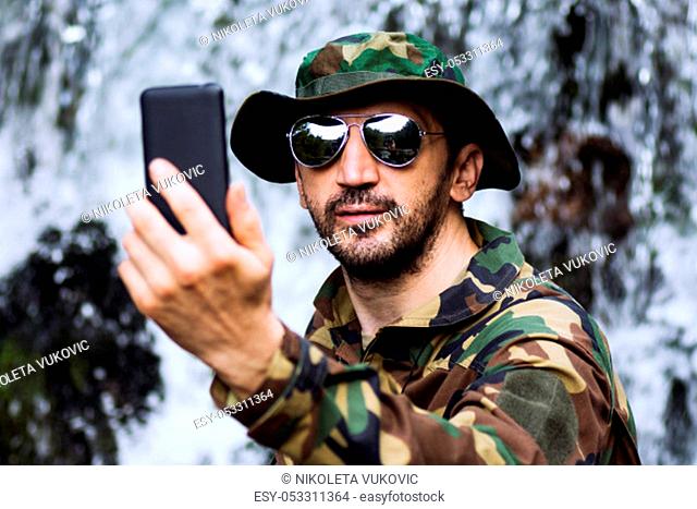 Man in military uniform is taking selfie and using phone in nature, adventure in nature