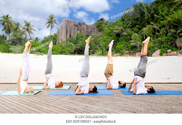 yoga, fitness, sport, and healthy lifestyle concept - group of people making supported shoulderstand pose on mat outdoors over exotic tropical beach background