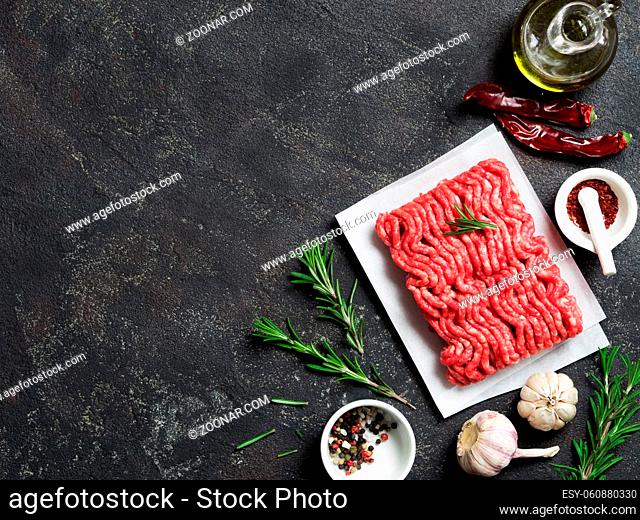 Fresh raw minced beef on backing paper and cutting board and ingredients over black cement background with copy space. Top view or flat-lay