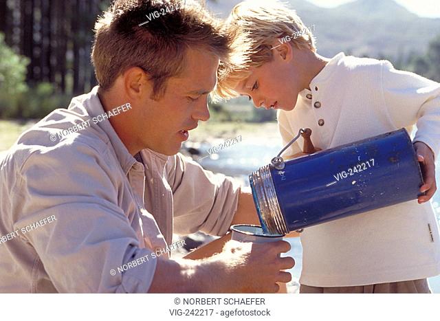 outdoor, close-up, blond 6-year-old boy holds a blue jug pouring something to drink into his father' s cup sitting at the shore of a lake in a forest  - GERMANY