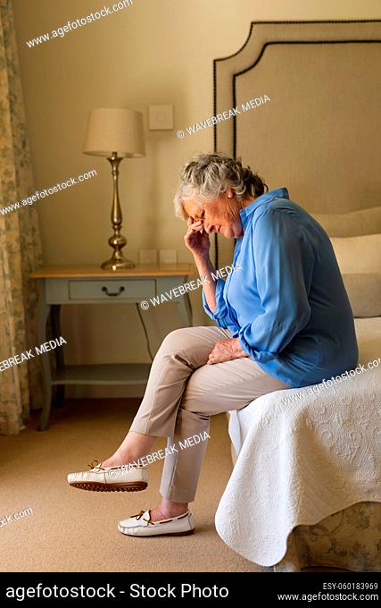 Senior caucasian woman sitting on bed holding her head and thinking in bedroom