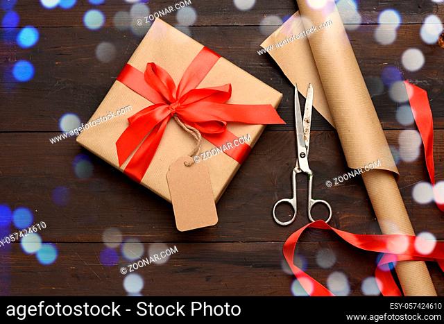 roll of brown kraft packaging paper, red silk ribbon, scissors and a bobbin with ribbon on a brown wooden background, top view