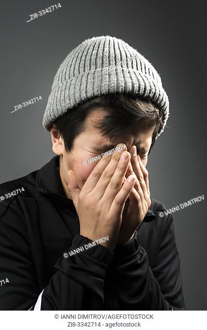 Young boy, 11 years old in black tracksuit and grey wooly hat crying with his face in his hands