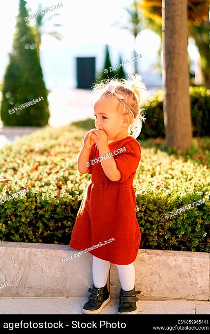 Pretty little girl in a terracotta colored dress and white tights is snacking on a pie in a park on a sunny day. High quality photo
