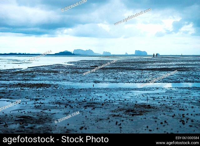 Couple walking in low tide in ocean with carst mountains in background and cloudscape at Koh Mook, Thailand, Asia