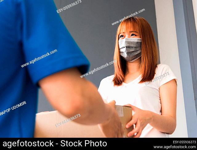 Asian young delivery man courier with package post box in uniform he protective face mask he making service woman customer receiving front house under curfew...
