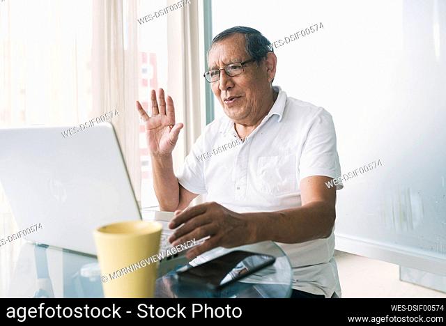 Senior man waving hand on video call through laptop at home