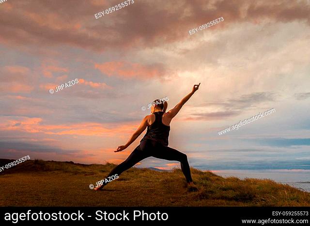 Middle-aged woman in black sportswear on her back doing meditation in front of the sea at sunset. Caucasian woman in yoga pose at dusk