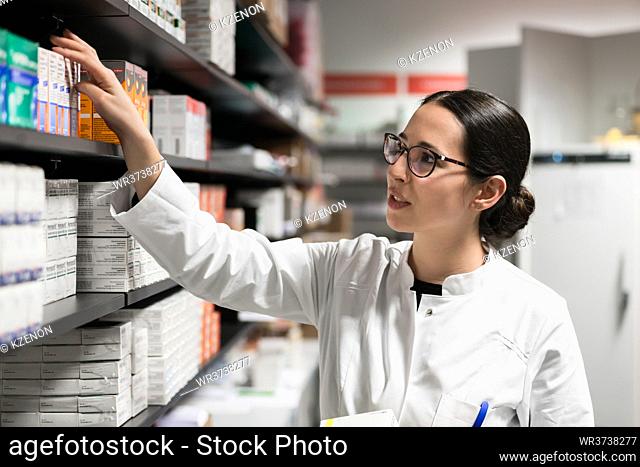 Portrait of a dedicated female pharmacist taking a medicine from the shelf, while wearing eyeglasses and lab coat during work in a modern drugstore with various...