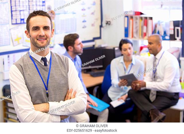 School teachers gather in a small school office for a chat. They look happy. The focus is on the man who stands in the foreground with the others sitting out of...