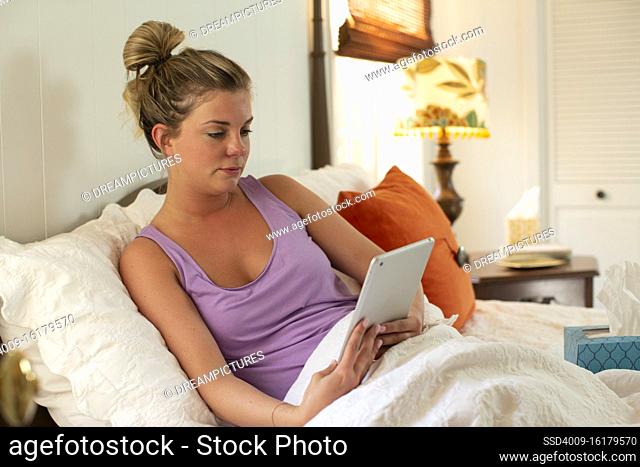 Young woman in her bed with box of tissue debating getting out of bed, using tablet to check in with doctor