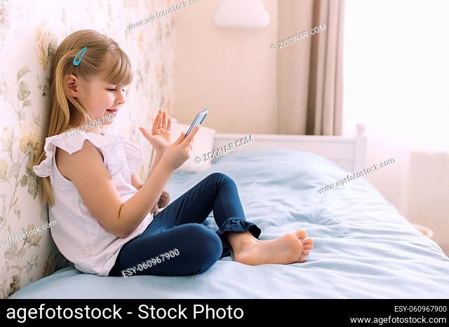 A little girl sits on the bed in the stylish bedroom, holding phone and have a video call using smartphone, waving a hand