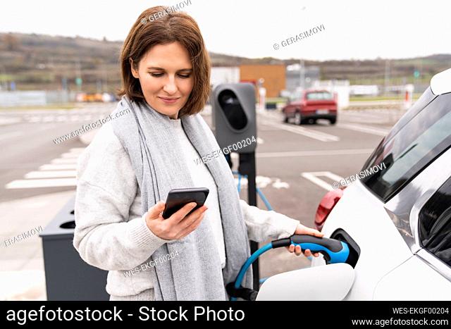 Woman using smart phone charging electric car at station
