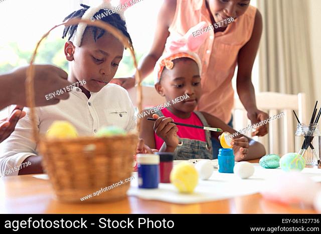 African american mother with son and daughter wearing bunny ears painting easter eggs