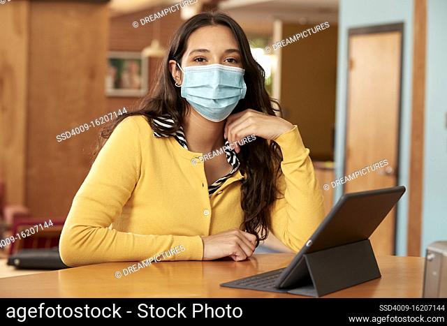 Portrait of young ethnic woman wearing yellow sweater with black and white striped blouse and face mask, sitting at bar in kitchen of downtown loft with iPad