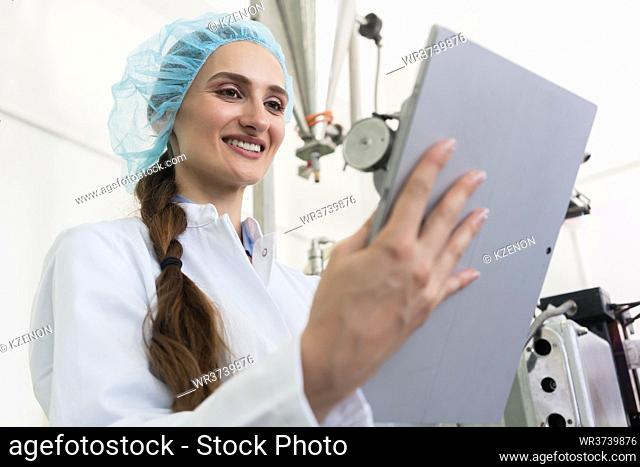 Female expert wearing white lab coat and protective headwear while analyzing information on a tablet PC during work in a contemporary factory