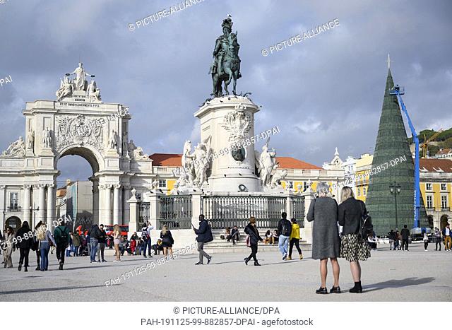 25 November 2019, Portugal, Lissabon: Tourists and street vendors stroll in the square of commerce in front of the triumphal arch of the Augusta street (l) and...