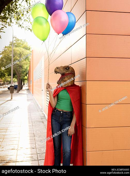 Young woman wearing crocodile mask standing with multi colored balloons by wall