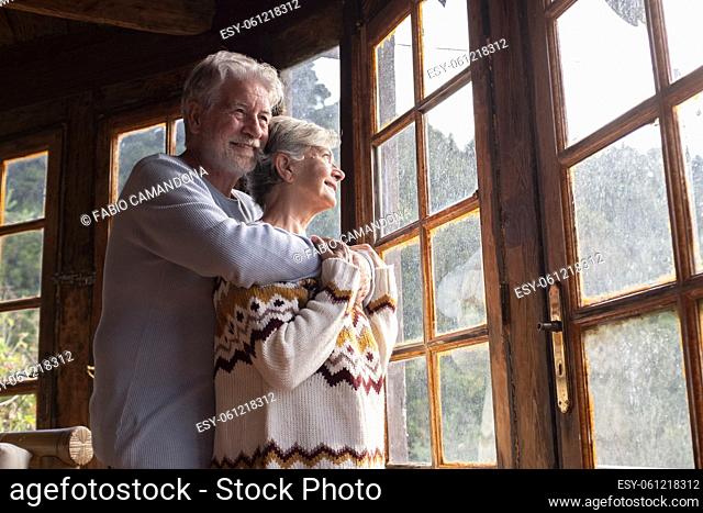 Elderly age relationship and love concept with couple of mature man and woman hugging and looking outside the windows at home with happiness and joy together