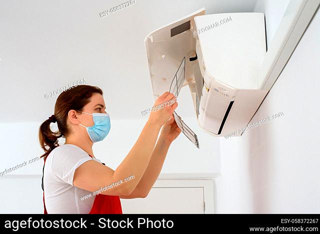 Female technician wearing face mask and cleaning air conditioner indoors
