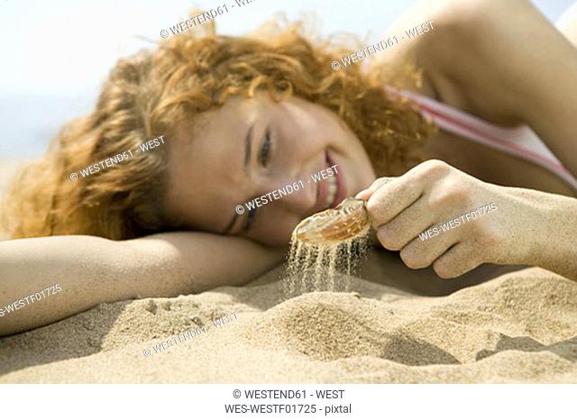 Young woman lying in sand, playing with shell, close-up
