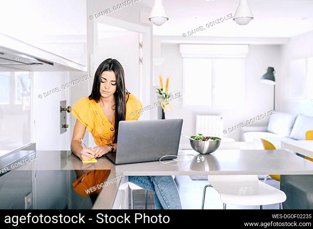 Young female freelance worker with laptop writing in notepad at home