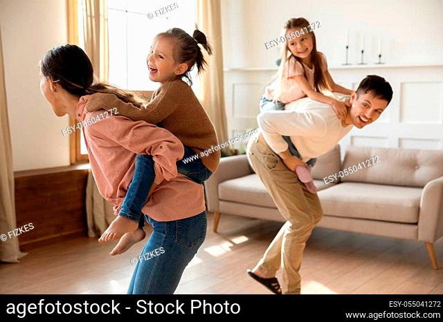 Happy parents with preschool children playing funny game, piggy back riding in living room, excited mother and father carrying two little daughters
