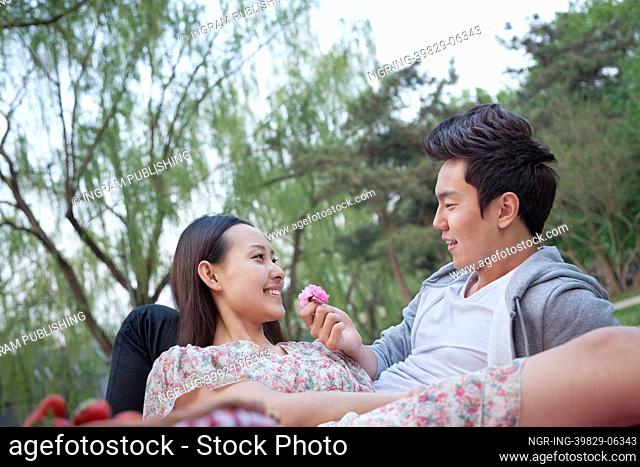 Smiling couple in love having a picnic in the park, lying down on the blanket and holding a flower blossom