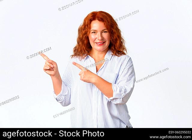 Smiling lovely middle-aged redhead woman showing announcement, pointing fingers upper left corner. Cheerful lady with ginger hair demontrate product banner over...