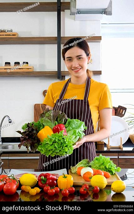 The housewife dressed in an apron holding a basket full of various kinds of vegetables on the front. Morning atmosphere in a modern kitchen