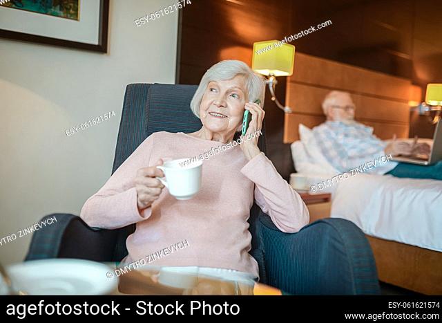 Morning. Senior good-looking lady having coffee and hoding a smartphone in hands