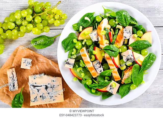 grilled chicken breast, grapes, spinach, gorgonzola cheese and apple salad on a white dish, on a table mat, on white wooden background, view from above