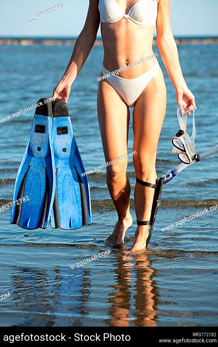 Young unrecognized woman wearing a white summer swimsuit and holding diving equipment at the beach. Sea activities