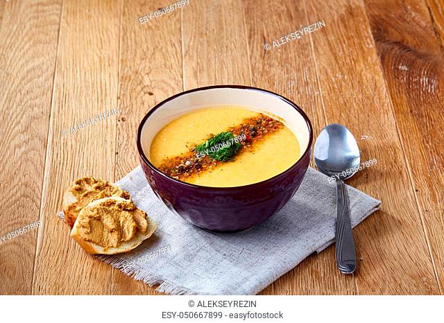 Purple bowl of creamy pumpkin soup garnished with dill, served with toasted bread slices covered with paste on homespun napkin over rustic background, closeup