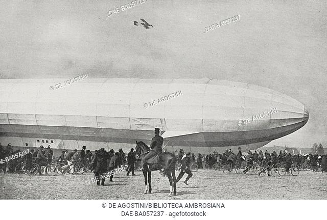 A large crowd watches a Zeppelin airship, while a French military biplane flies over the area, Luneville, France, photograph from the magazine L'Illustration