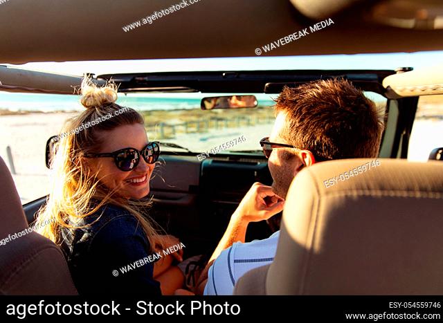 Rear view of a Caucasian couple sitting inside an open top car with blue sky and sea in the background, the man driving and the woman smiling to camera