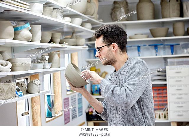 A man inspecting a clay pot, before firing. Shelves in a pottery studio full of pots, work in progress